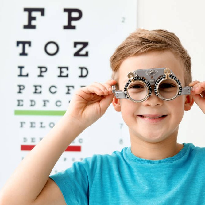 Little boy with trial frame near eye chart in ophthalmologist office