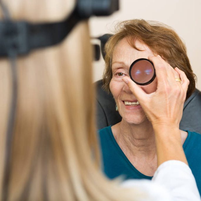 Female optician examining senior woman's eye with binocular indirect ophthalmoscope